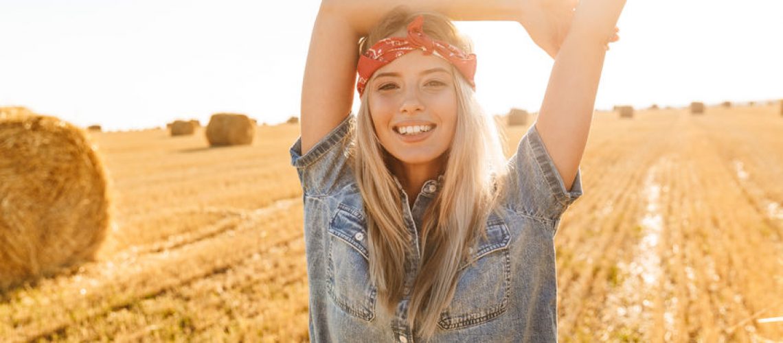 Portrait of countryside woman 20s smiling and walking through golden field during sunny day