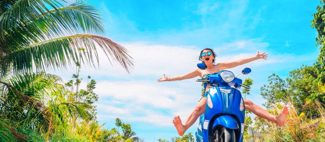 Crazy funny woman with flying hair riding a motorbike on a blue sky and green tropics background. Young girl with dark hair in sunglasses on a blue scooter in vintage style racing downhill. Concept of summer holidays and vacation adventures and wanderlust.