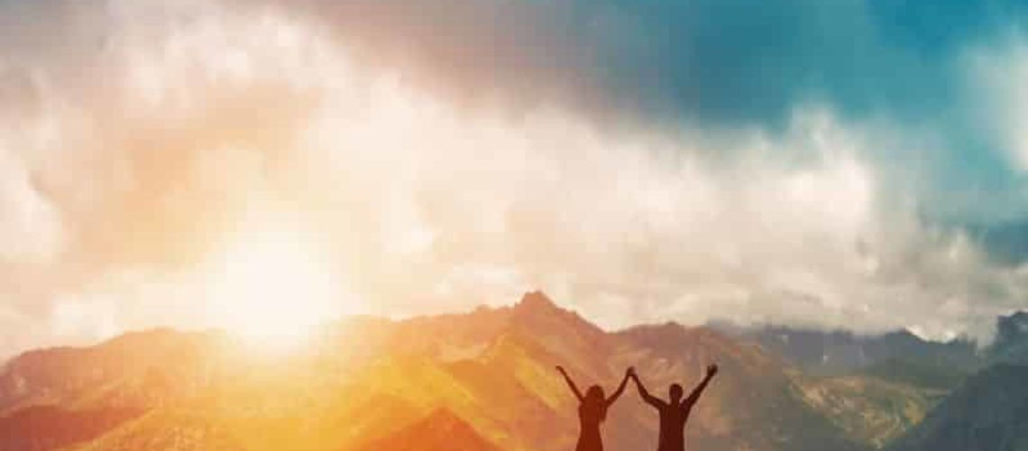 26507711 - a happy couple standing together on the peak of a mountain with hands raised admiring breathtaking view at sunset